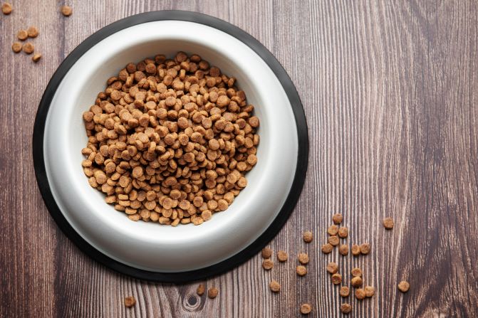 A photograph of a dog bowl filled with dry dog food on a laminate floor